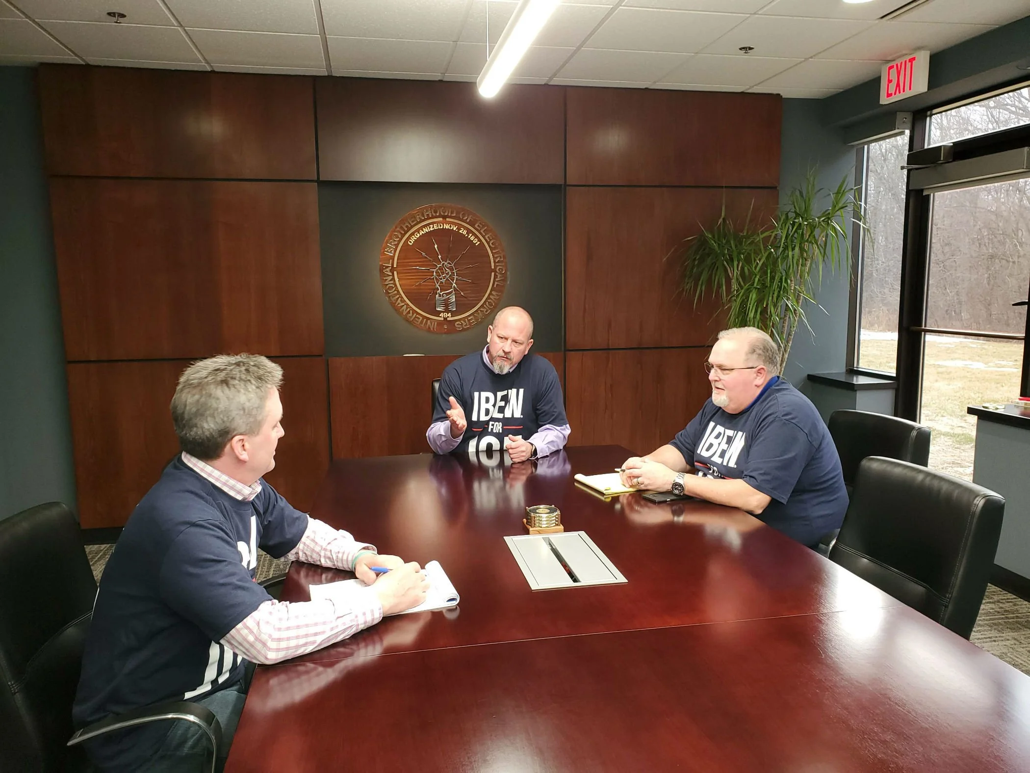 People sitting around a conference table engaged in discussion in a corporate office, with a wall emblem that reads 'International Brotherhood of Electrical Workers' behind them.