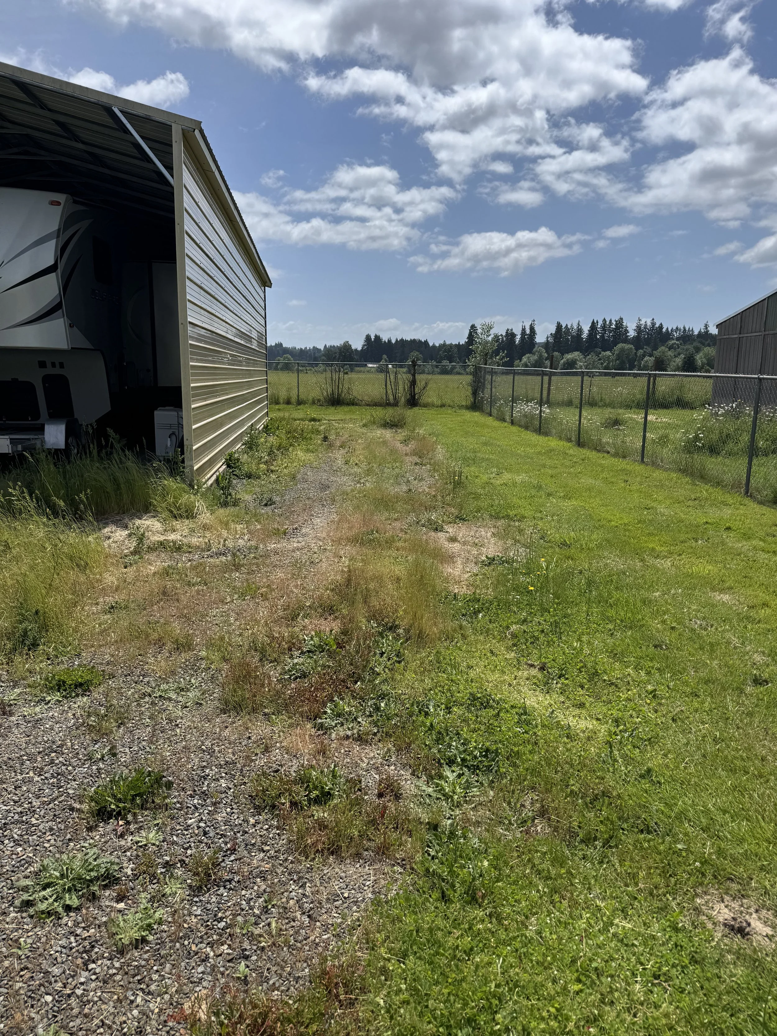 View of an outdoor grassy area with a paved path on the left side, a chain-link fence bordering a field, and a metal building on the right. The sky is partly cloudy with blue skies and white clouds.