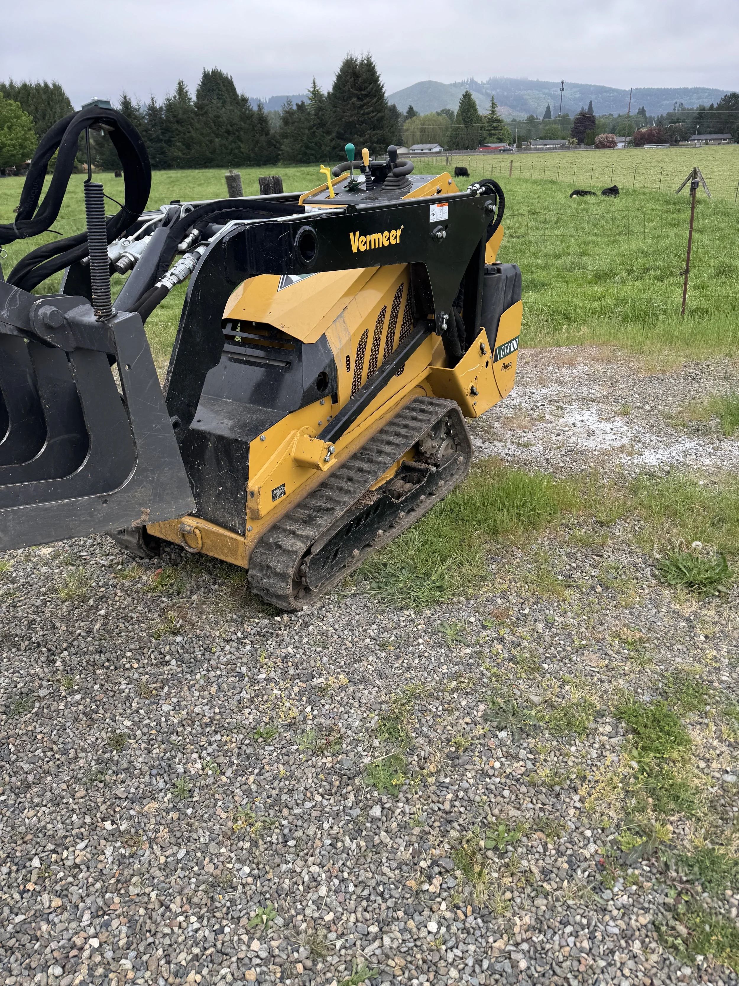 A small tracked construction vehicle with the brand name 'Vermeer' on it, parked on a gravel area near a grassy field with some cows and trees in the background under a cloudy sky.
