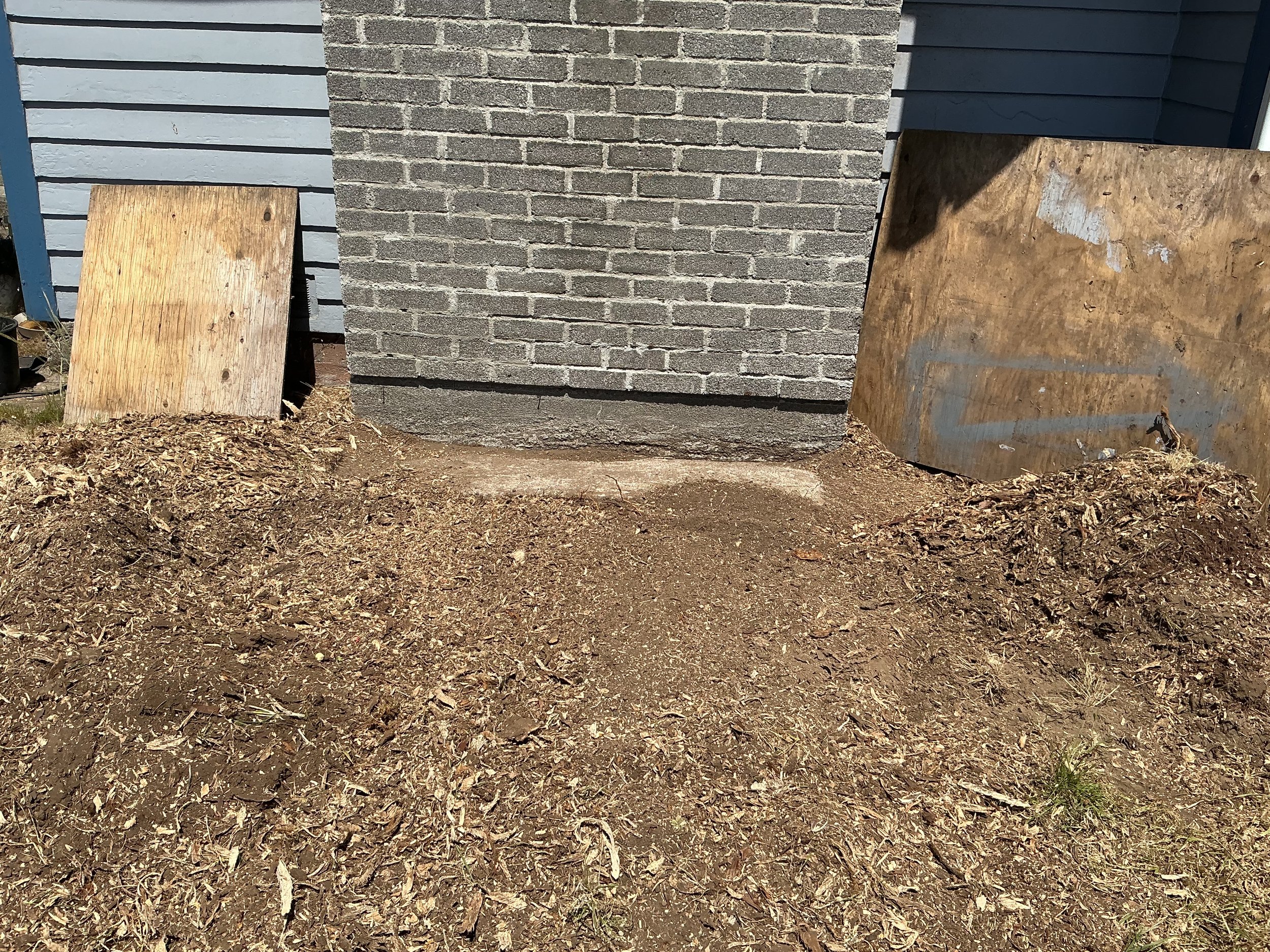 A brick wall with two wooden boards leaning against it on dirt ground covered with dry leaves.