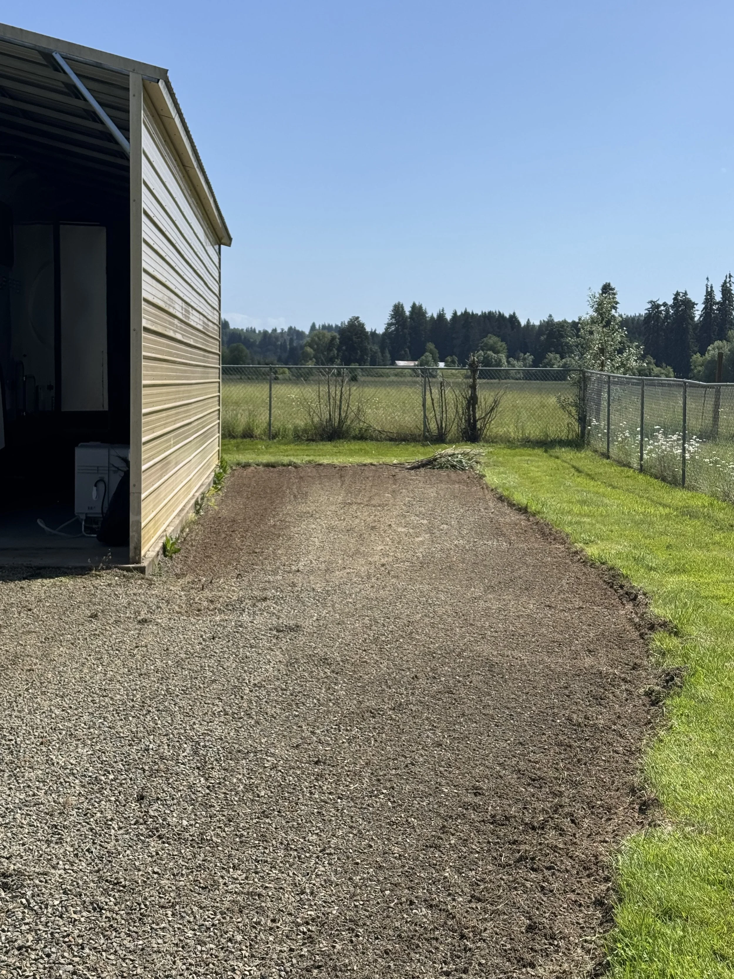 View of a gravel pathway running alongside a house with a metal exterior wall, fence, green grass, trees, and a clear blue sky.