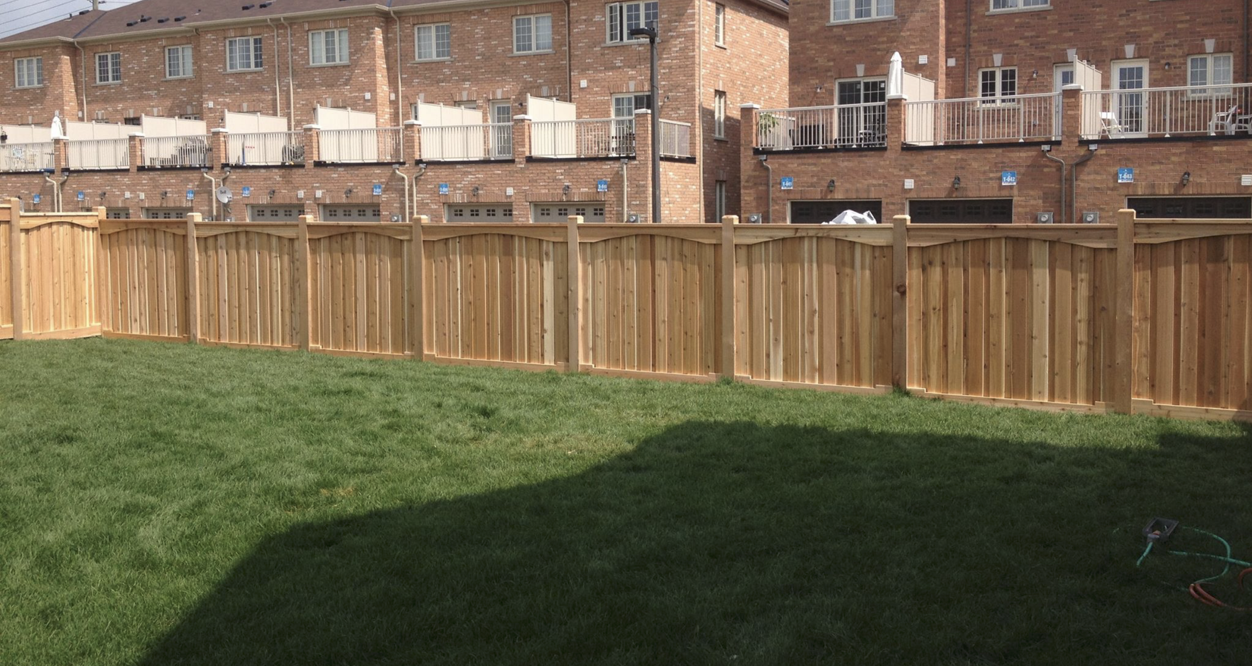 A backyard with a green lawn, a wooden privacy fence, and a brick apartment building in the background.