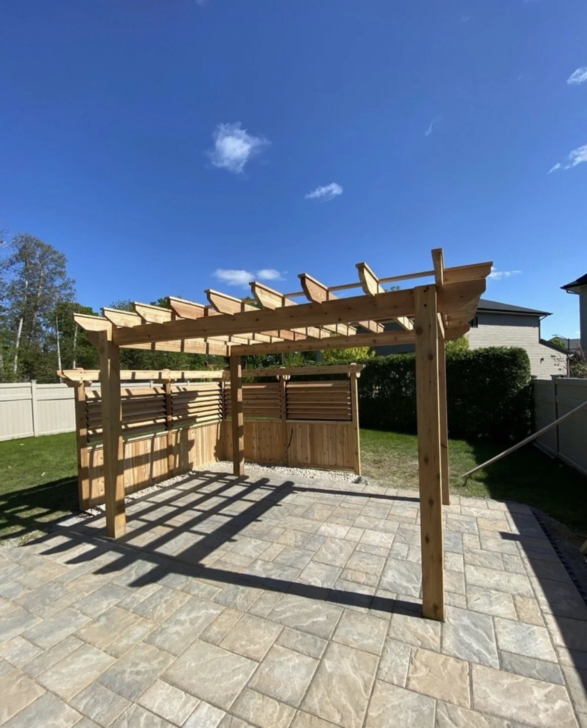 Wooden pergola under construction in a backyard with stone patio, surrounded by grass, trees, and a white fence.