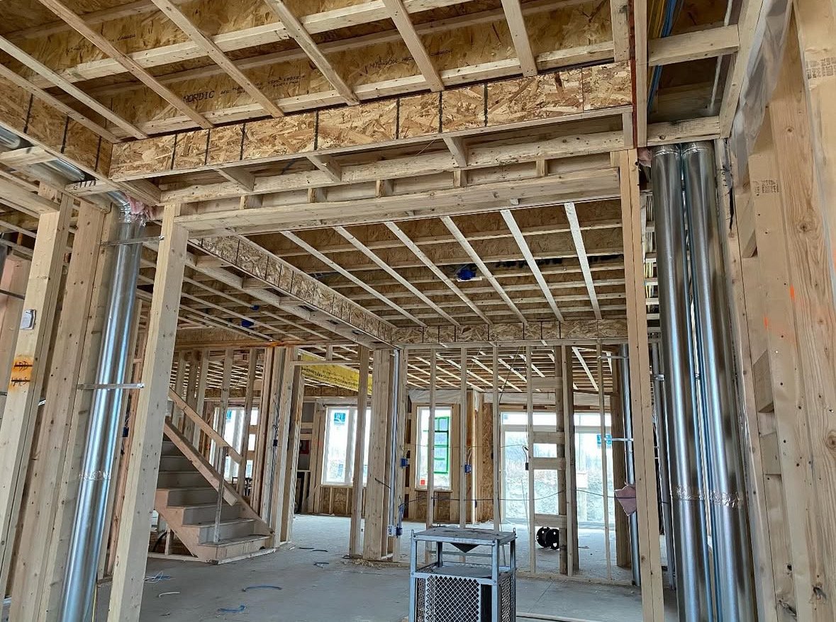 The interior of a house under construction showing wooden framing, metal pipes, and a staircase leading to an upper floor with windows in the background.