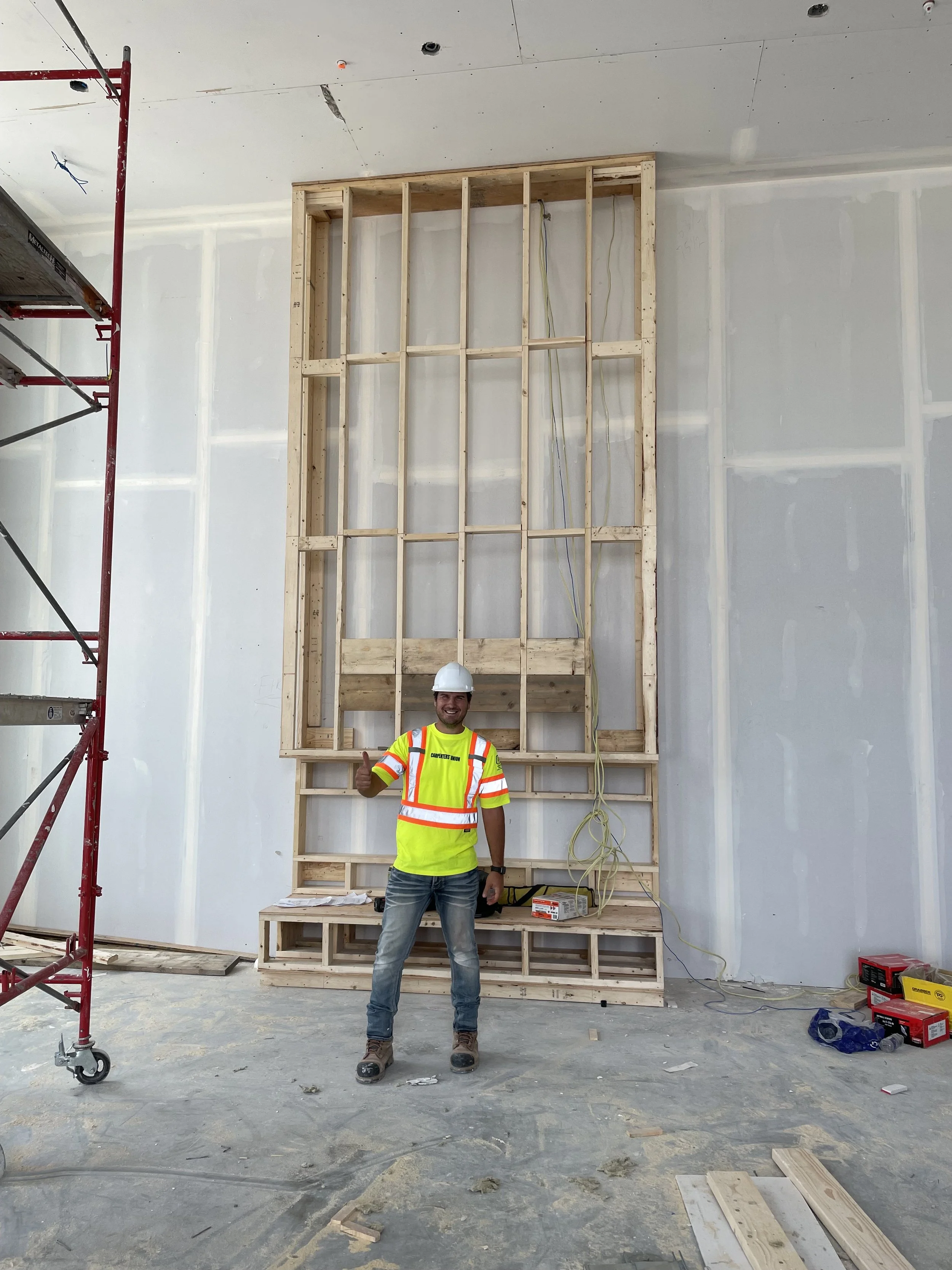A man wearing a yellow construction safety vest, jeans, work boots, and a white hard hat, standing inside a building under construction. He is smiling and giving a thumbs-up in front of a wooden framed wall, with construction tools and materials scat