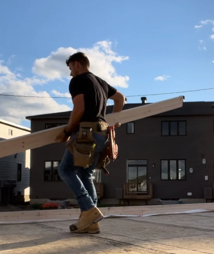 A construction worker walking on a wooden platform carrying a plank, wearing a tool belt and work boots, with a modern house and a blue sky with clouds in the background.