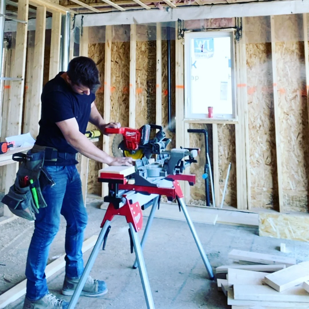 A man working at a construction site inside a room with unfinished walls. He is using a saw on a piece of wood, with various tools hanging from his tool belt.