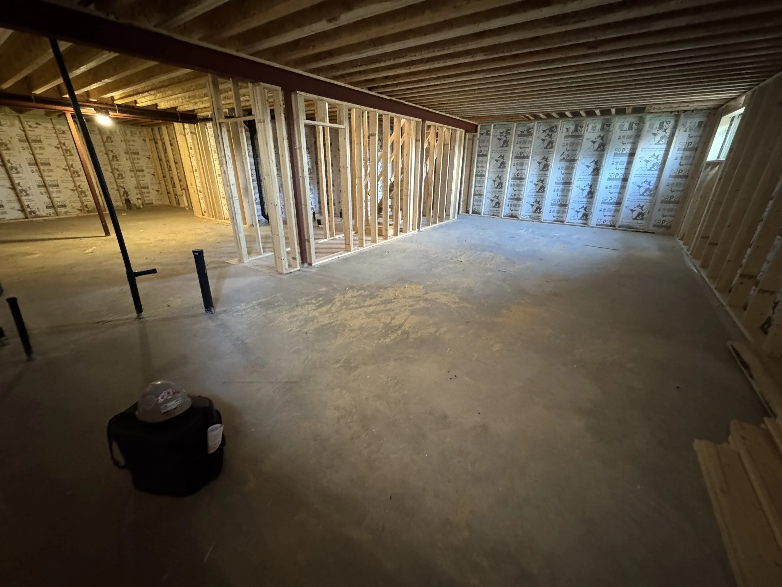 Interior view of a house under construction with unfinished framed walls, exposed ceiling beams, and a concrete floor. Construction tools and a helmet are visible in the foreground.