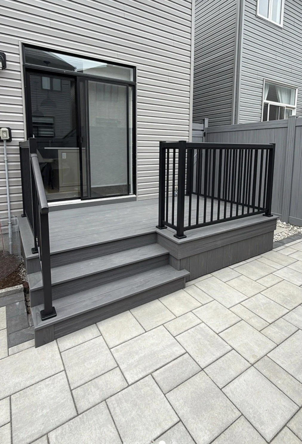Backyard patio with steps, gray deck, black railing, sliding glass door, and surrounding gray fencing.