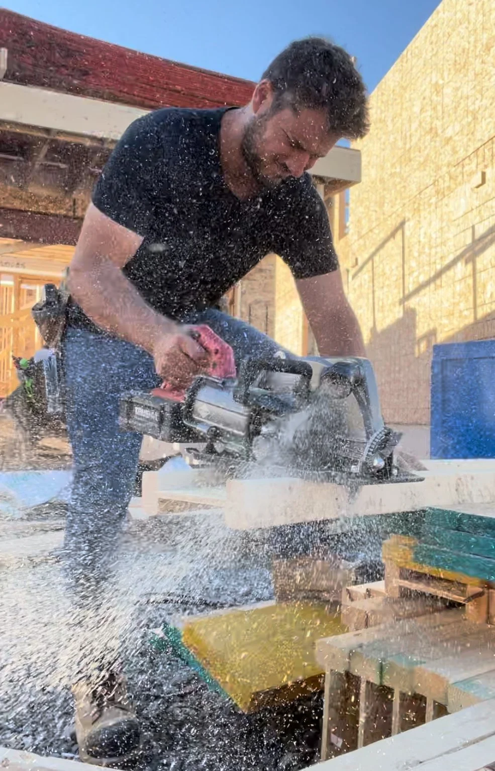 A man using a handheld power saw to cut wood outdoors, with sawdust and debris flying in the air.
