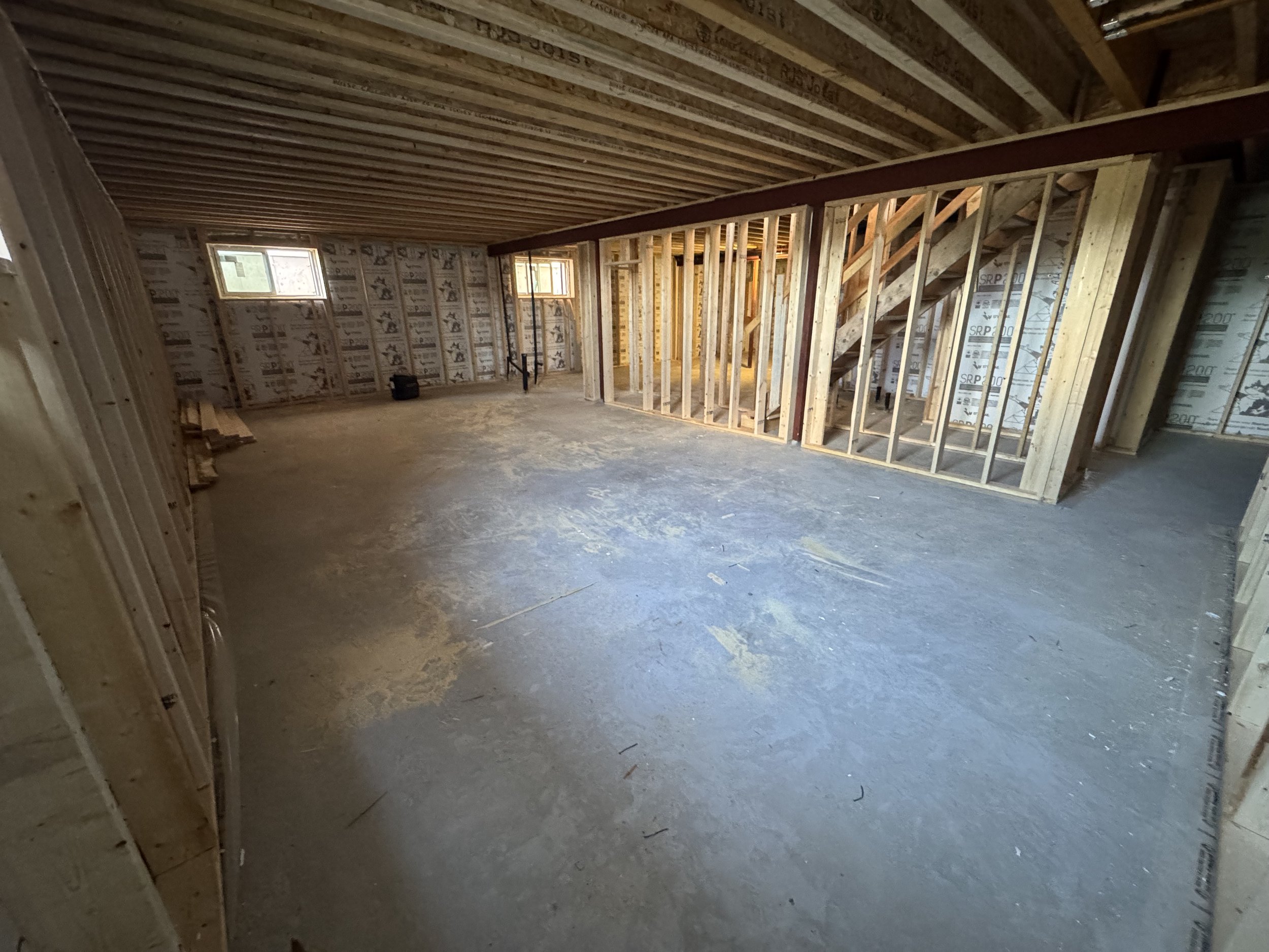 Interior view of a house under construction, showing exposed wooden framing, stairs, and small windows, with a concrete floor.