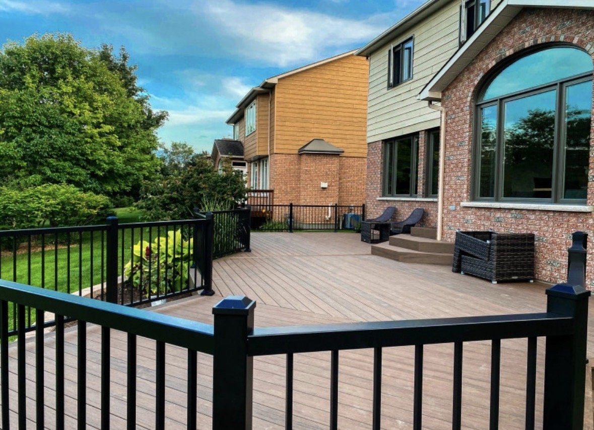 A spacious wooden deck attached to a brick house with large arched windows, outdoor seating, and a black metal railing, overlooking a lush green yard and trees under a partly cloudy sky.