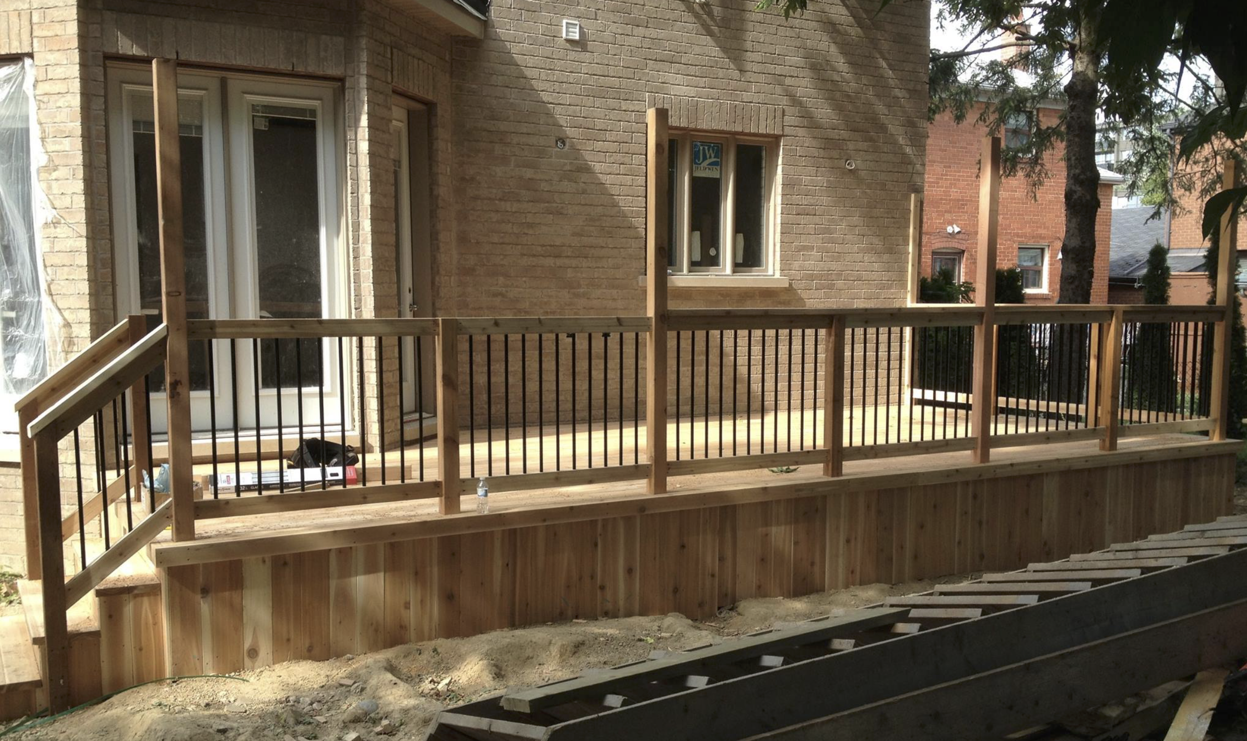 Backyard construction site with a wooden deck and railing, brick house exterior, windows, trees, and building materials.