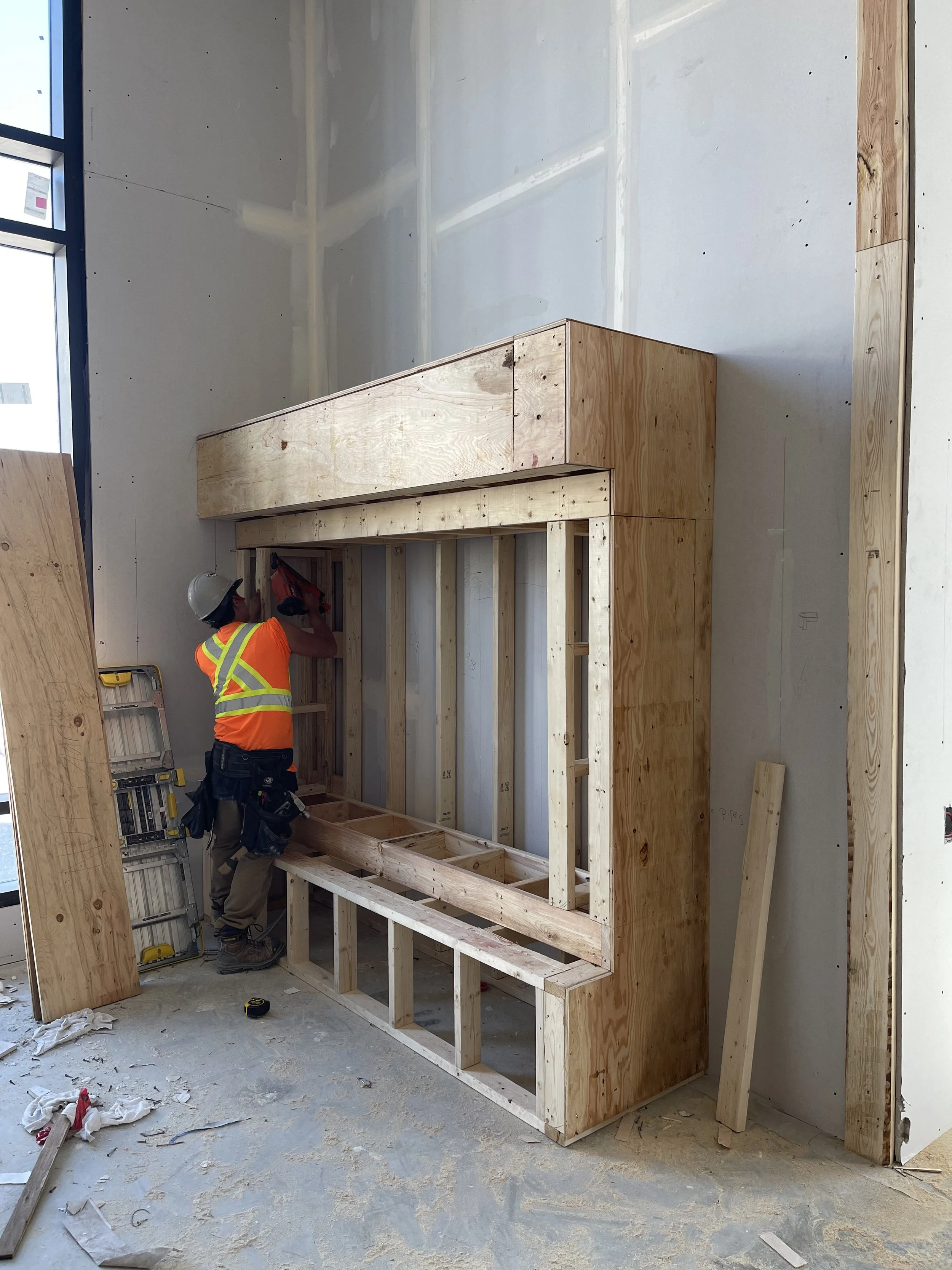 Construction site with a worker in a safety vest and helmet building a wooden frame structure against a drywall wall, surrounded by tools and materials on the floor.