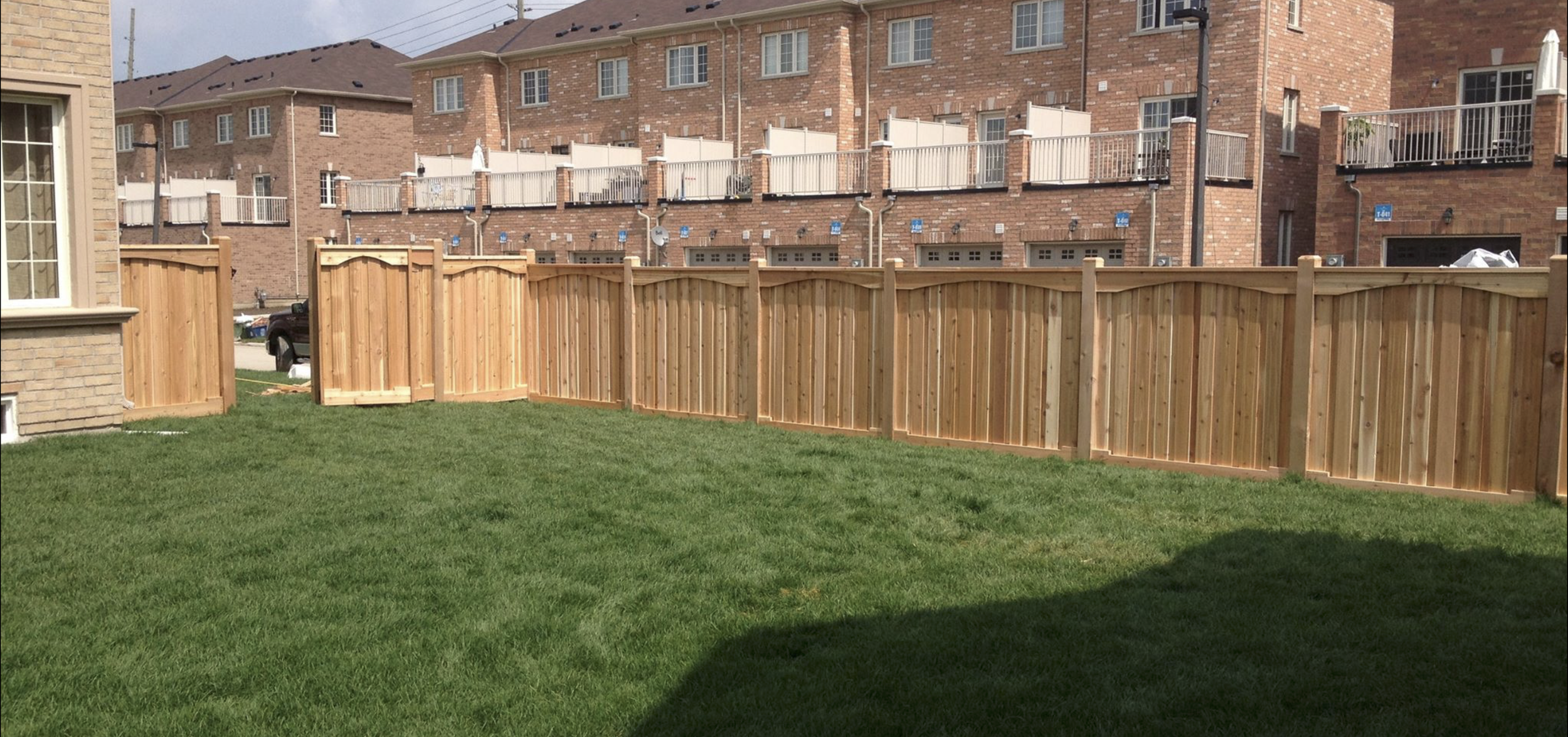 View of a backyard with a wooden fence, green grass, and brick apartment buildings in the background.
