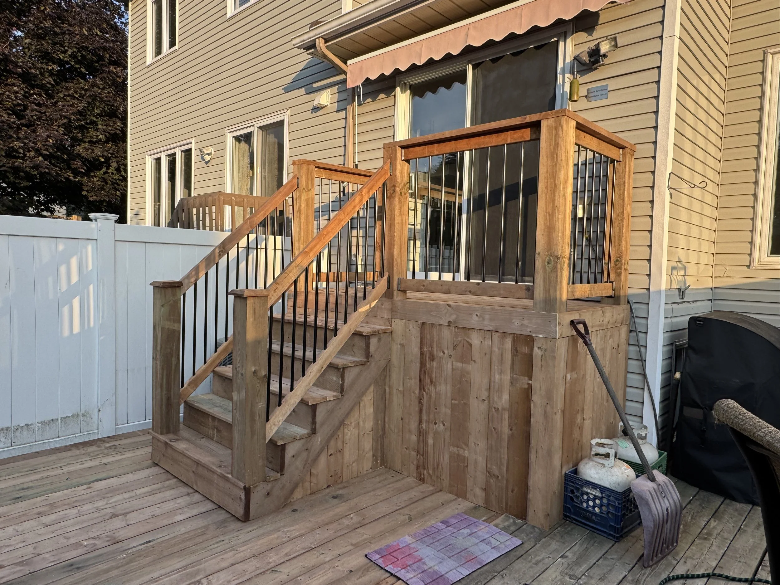 Edited image of a house's backyard showing new wooden stairs leading to a small deck with a railing, adjacent to a white fence, with a patio mat and construction tools visible.