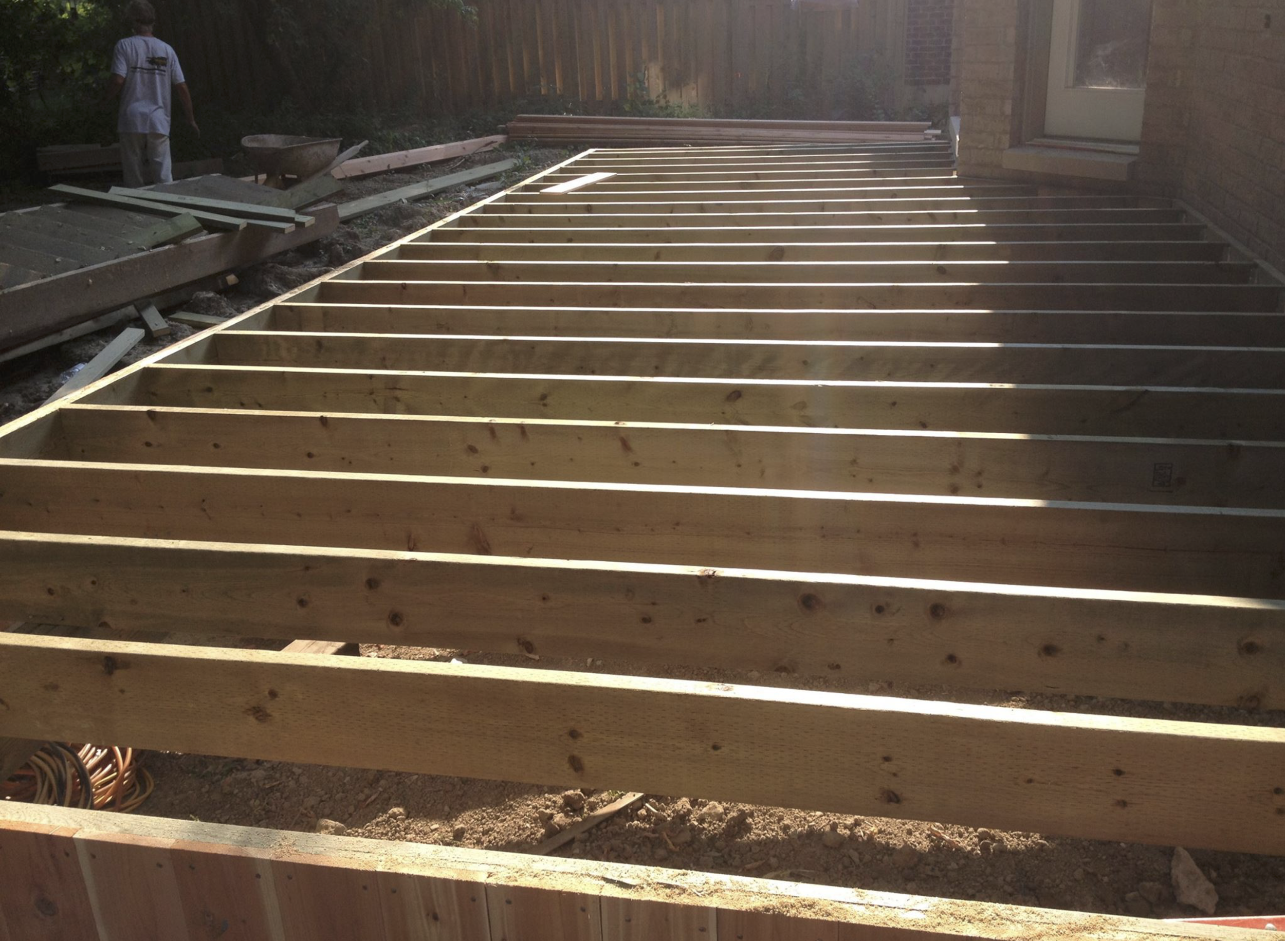 Partial view of a wooden deck under construction next to a brick house, with a person working in the background.