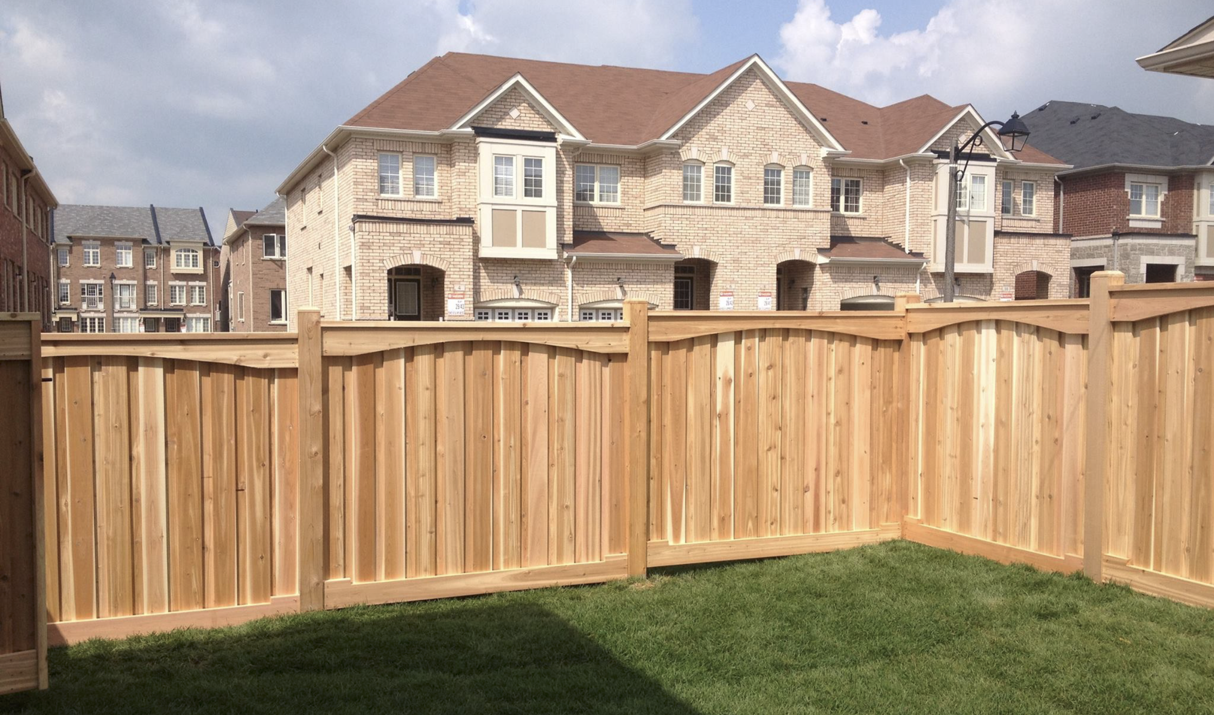 A wooden privacy fence in front of a row of brick and stucco houses with brown and gray roofs, under a partly cloudy sky.