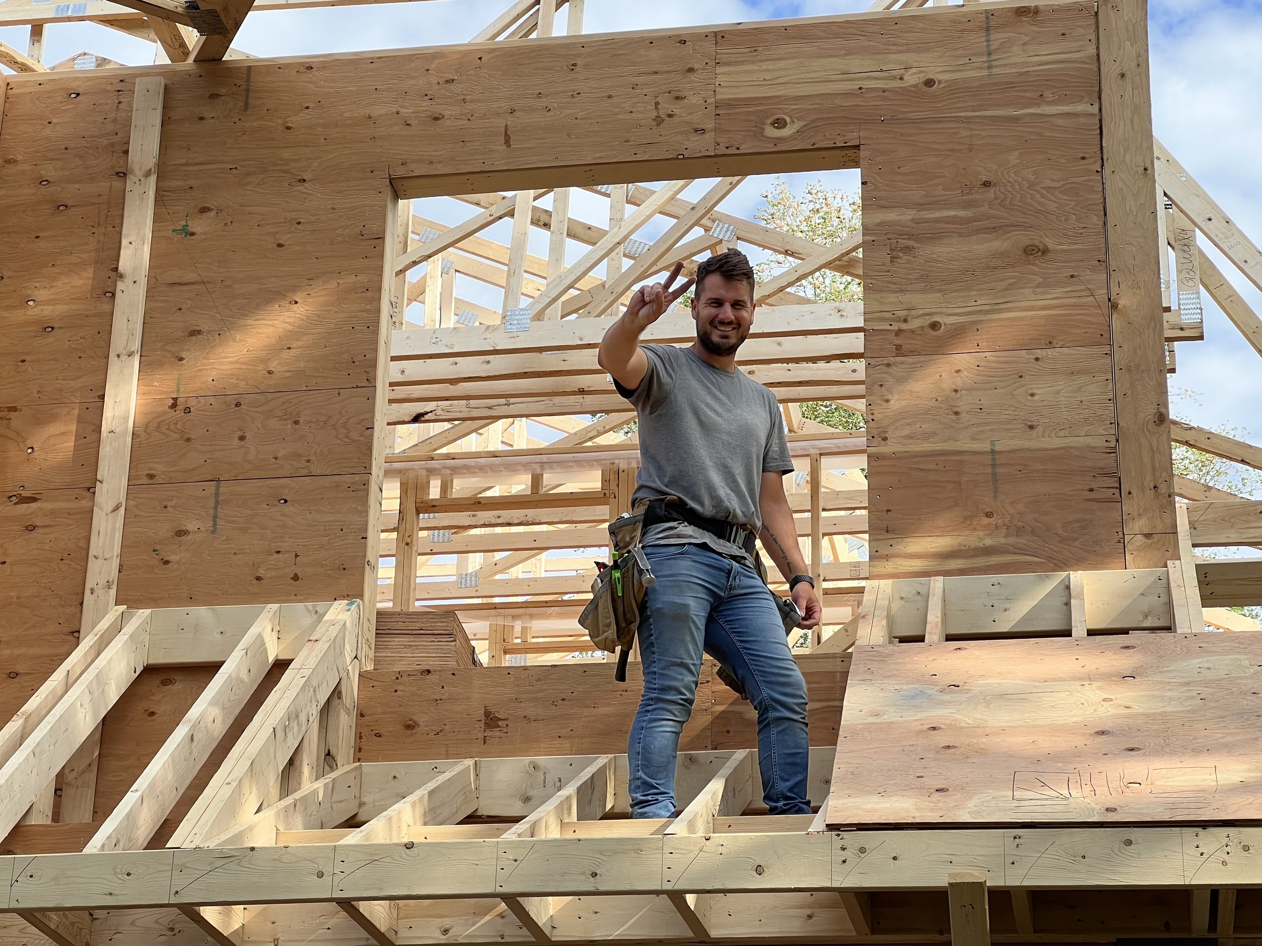Construction site with wooden framing, a smiling man in a gray t-shirt making a peace sign, standing inside the structure.