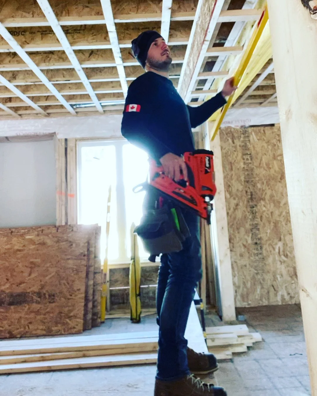 A construction worker in a black shirt with a Canadian flag patch, a black beanie, and jeans, is standing inside a building under construction. He is holding a yellow level against the ceiling framework and has a red power tool attached to his belt. There are wooden planks and a ladder in the background, with sunlight coming through an unfinished window.