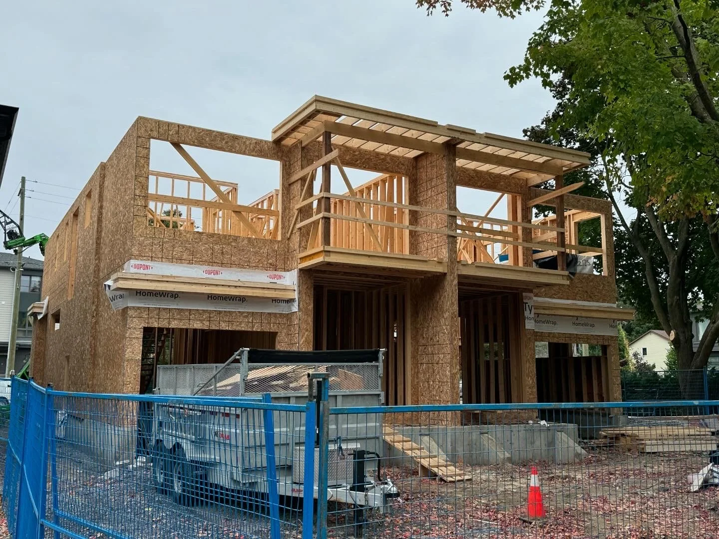 Wood frame house under construction with two stories, wooden beams, and plywood sheathing, surrounded by construction fencing and equipment.