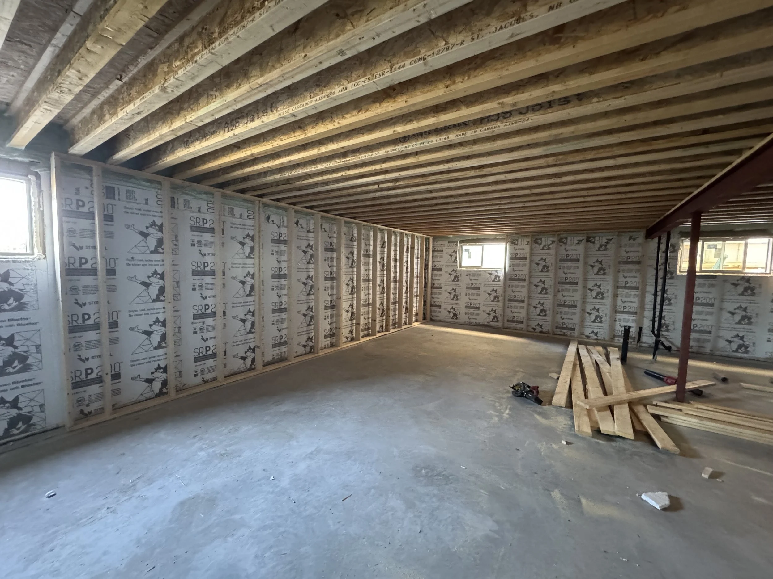 Interior of a building under construction with exposed wooden ceiling beams and partially installed wall insulation. Construction tools and materials are on the floor.