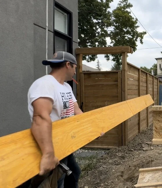 Man wearing a gray and white baseball cap and a white T-shirt holding a large wooden beam near a construction site with a gray building, wooden fence, and trees in the background.