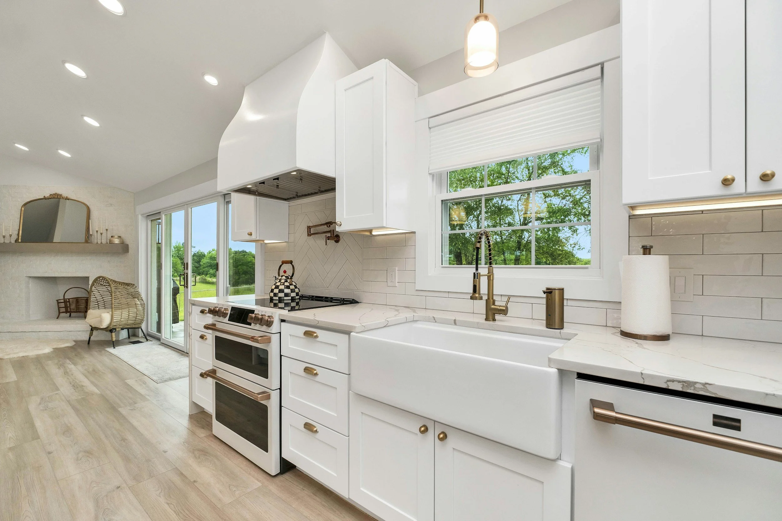 Modern kitchen with white cabinets, marble countertop, gold hardware, large window with outdoor greenery, and a view of the living area with a fireplace and armchair.