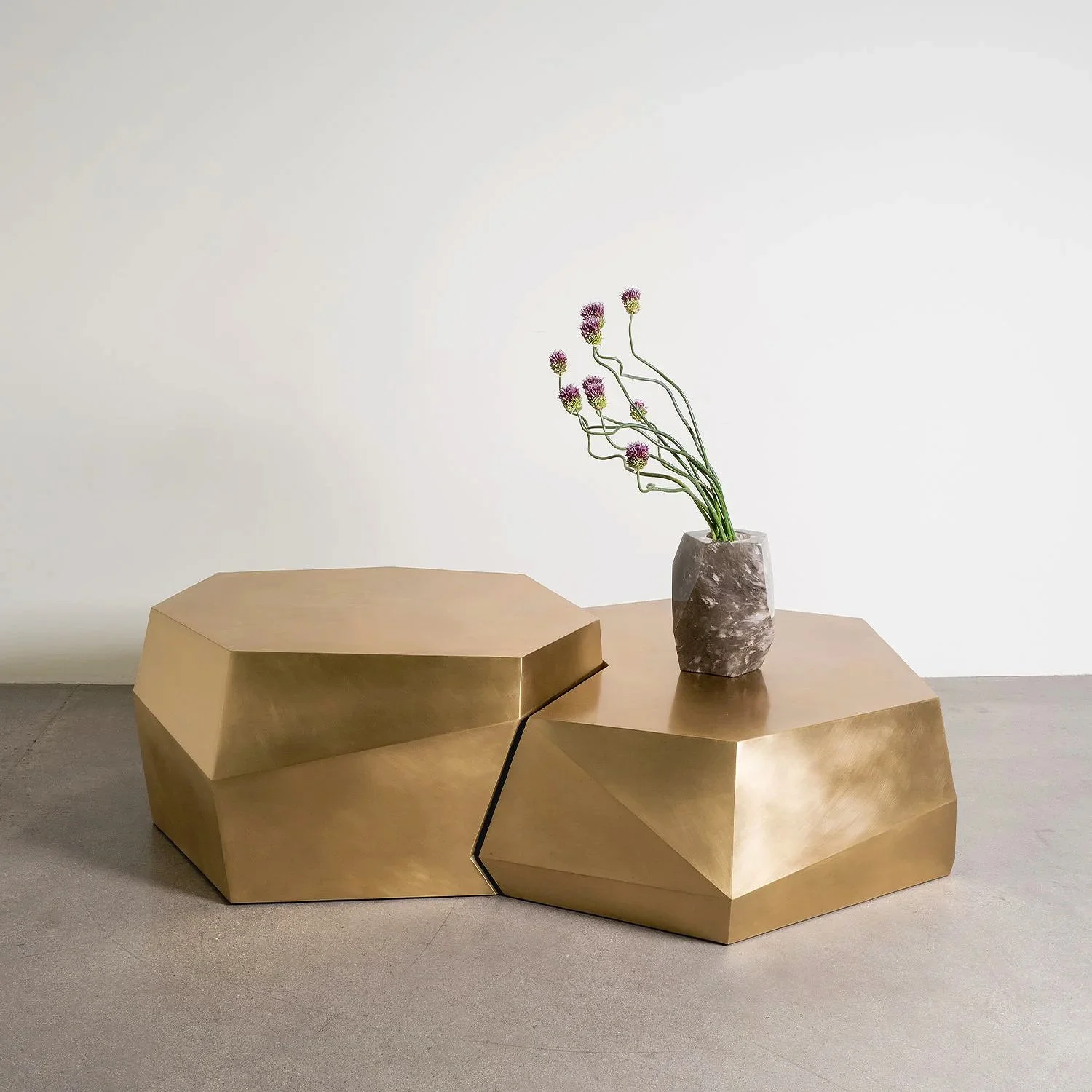 A modern, angular gold-colored table with a marble vase holding purple and pink flowers placed on top, against a plain white wall.