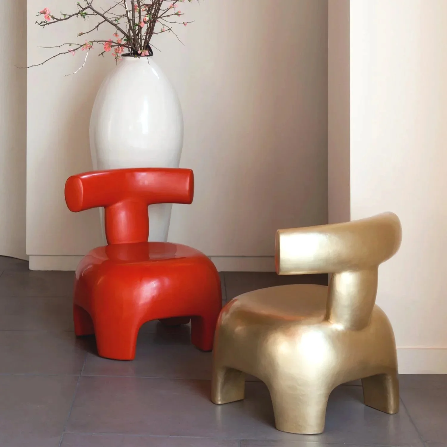 Decorative interior scene with a large white vase with pink blossoms and branches, a red sculptural chair, and a gold sculptural chair, all on a tiled floor against a plain wall.