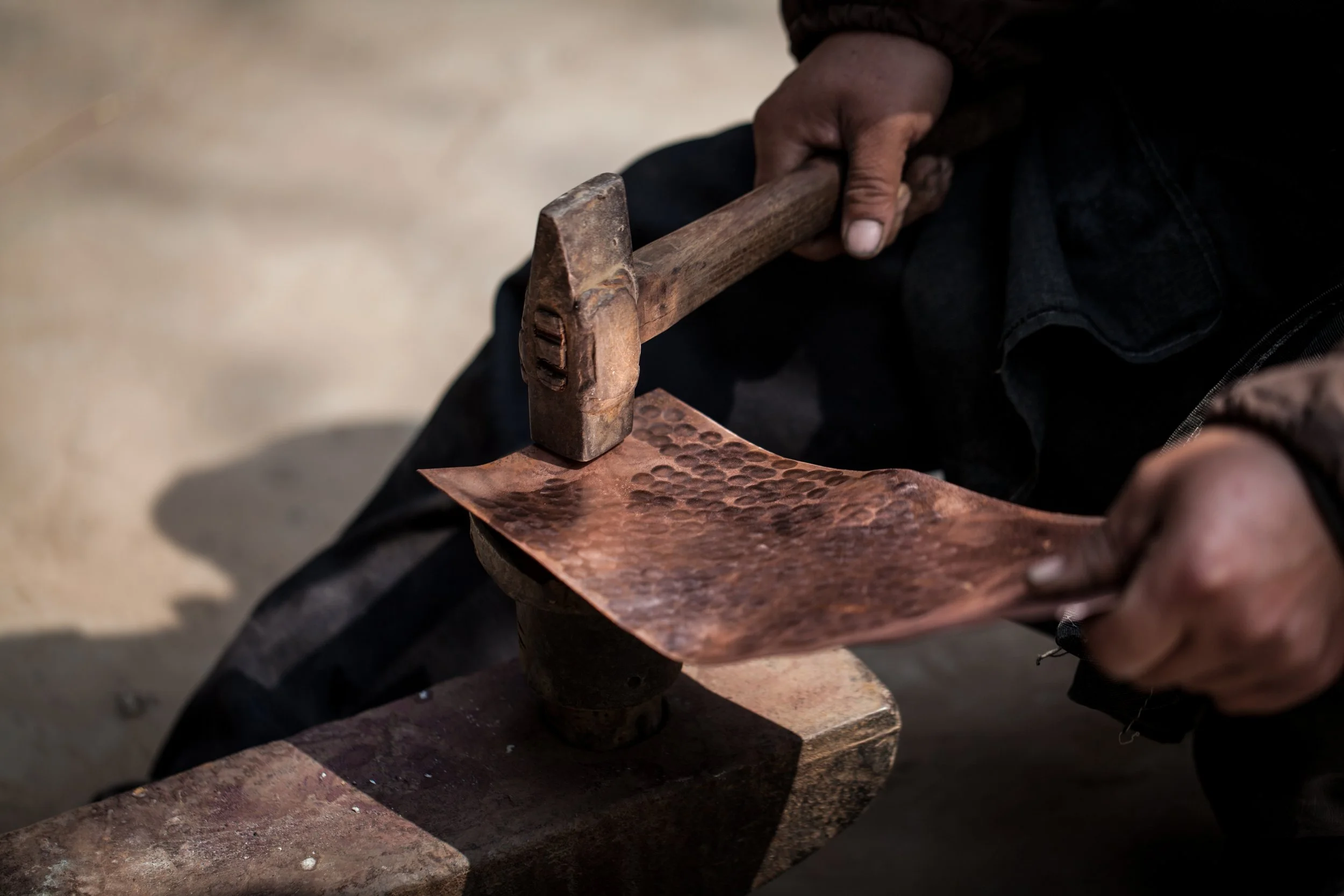 Close-up of a person's hand holding a hammer above a hammered copper sheet.