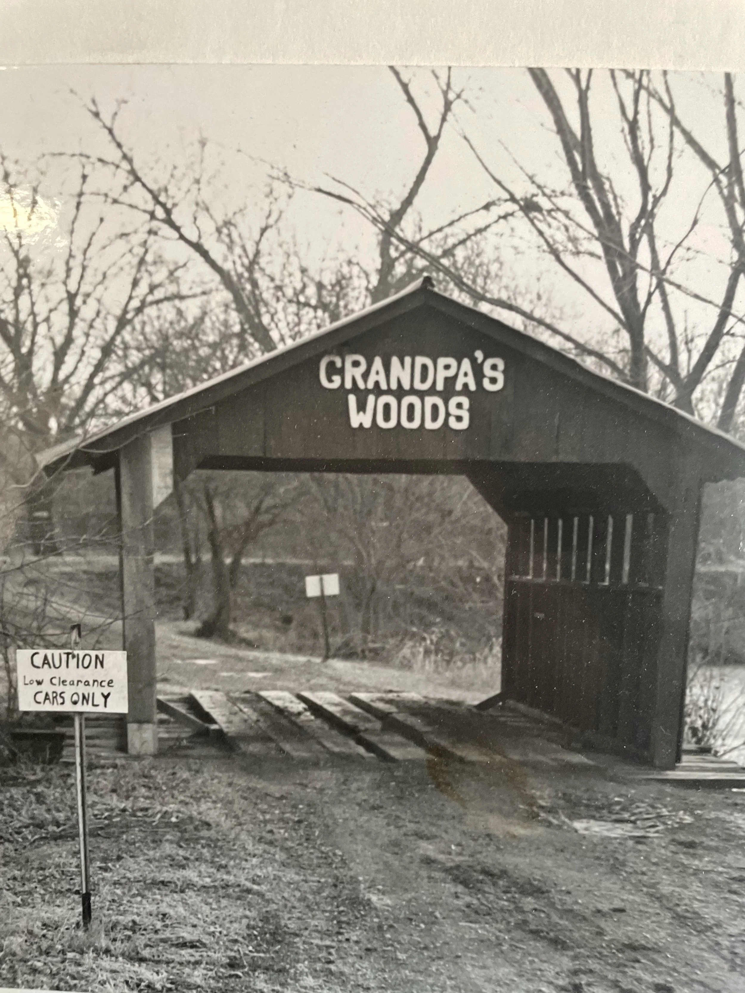 Black and white photo of a park entrance with a sign that says 'Grandpa's Woods' at the top. There is a smaller sign warning 'Caution low clearance cars only.' Tree branches are visible overhead, and the ground is covered with leaves and gravel.