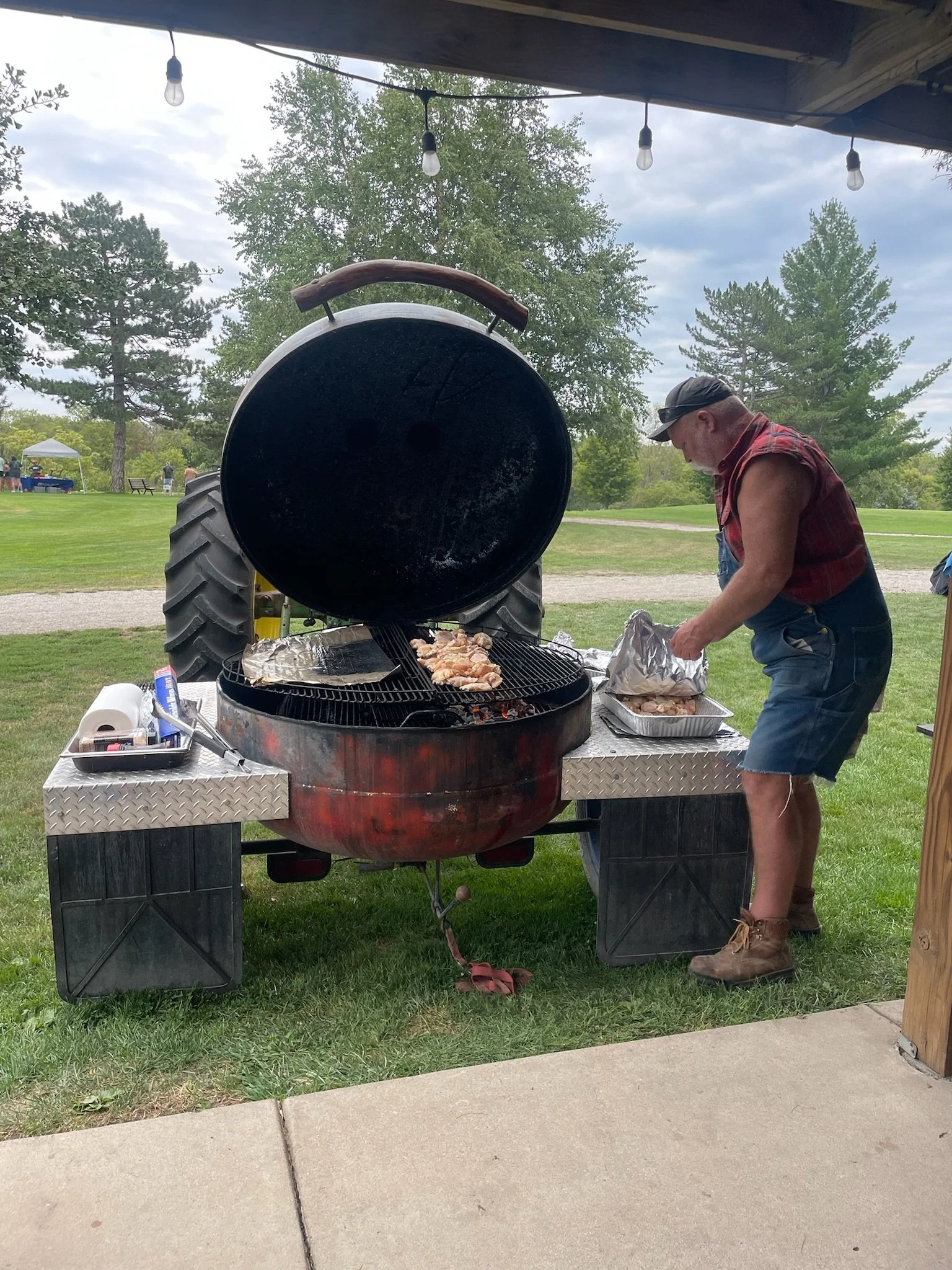 A man wearing a sleeveless plaid shirt, shorts, and work boots stands by a large outdoor grill, cooking meat. The grill is round with a large lid open, and the scene is outdoors with trees and a cloudy sky in the background.