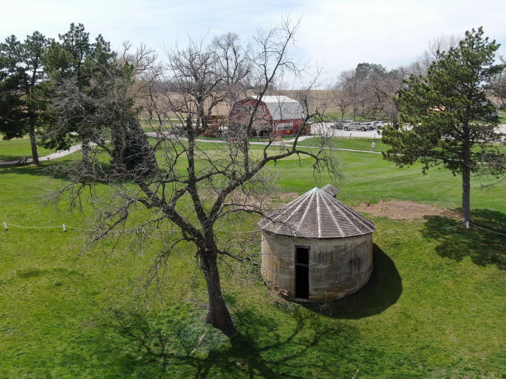 A park with green grass, several trees with bare branches and some with green pine needles, a small circular building with a conical roof, a red barn, parked cars, and a person walking along a paved path under a partly cloudy sky.