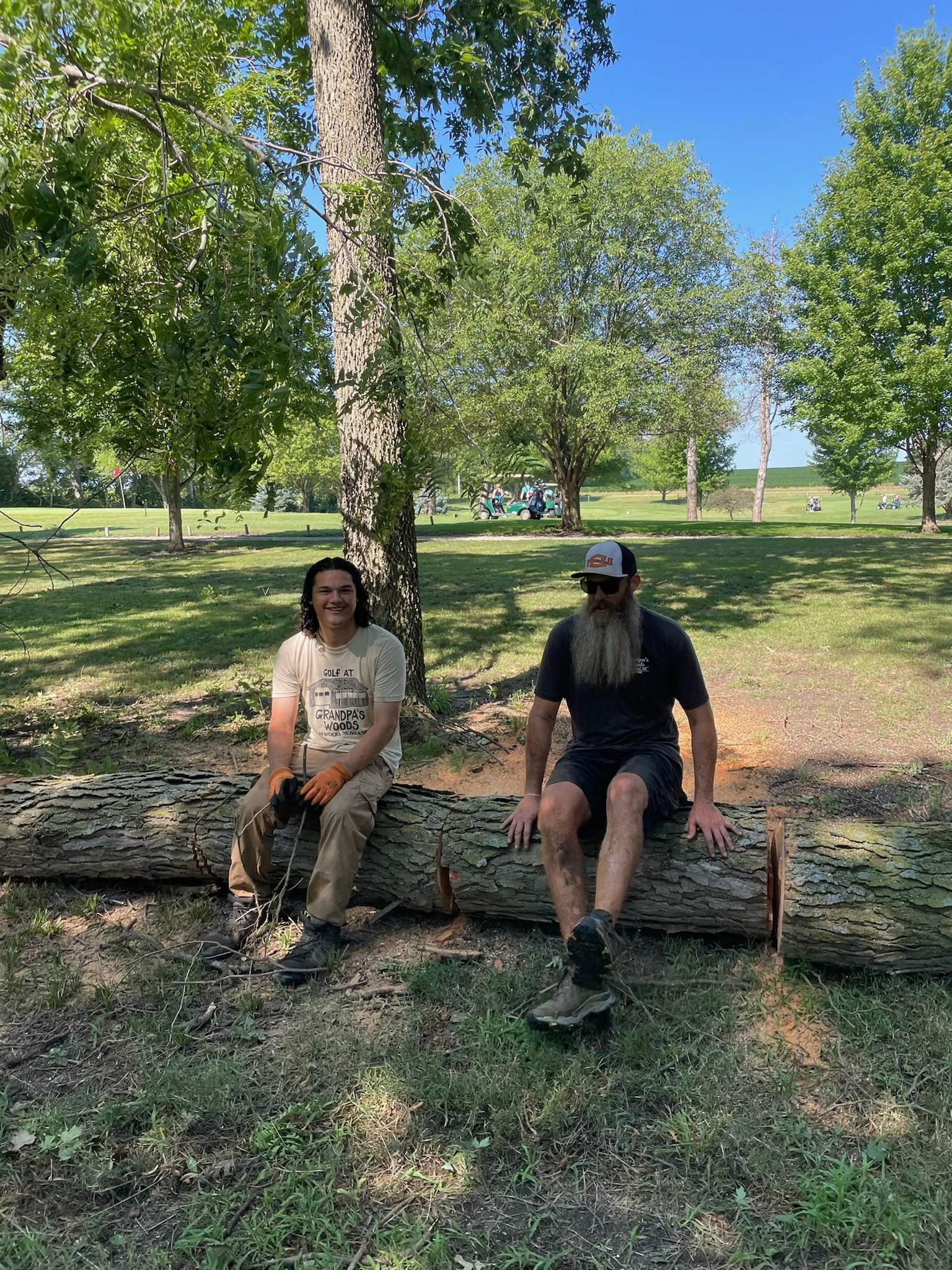 Two men sitting on a fallen tree trunk in a park with green trees and a golf course in the background. One man has long hair and is smiling, wearing a beige t-shirt and orange gloves. The other man has a long beard, sunglasses, a cap, and a black t-shirt. It is a sunny day with a clear blue sky.