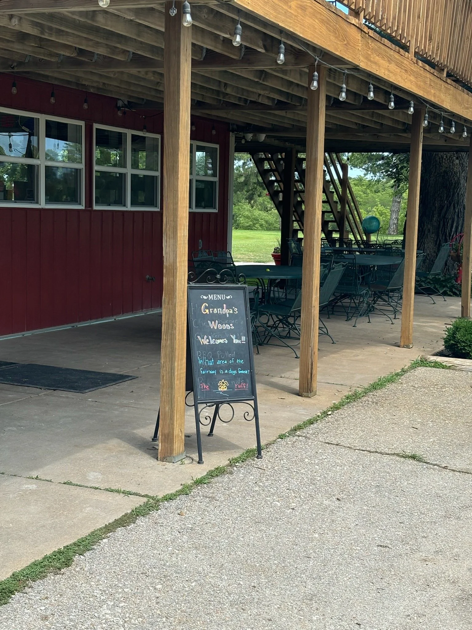 Outdoor patio area with black metal tables and chairs, a chalkboard menu, and a wooden deck with string lights. There are trees and greenery in the background.