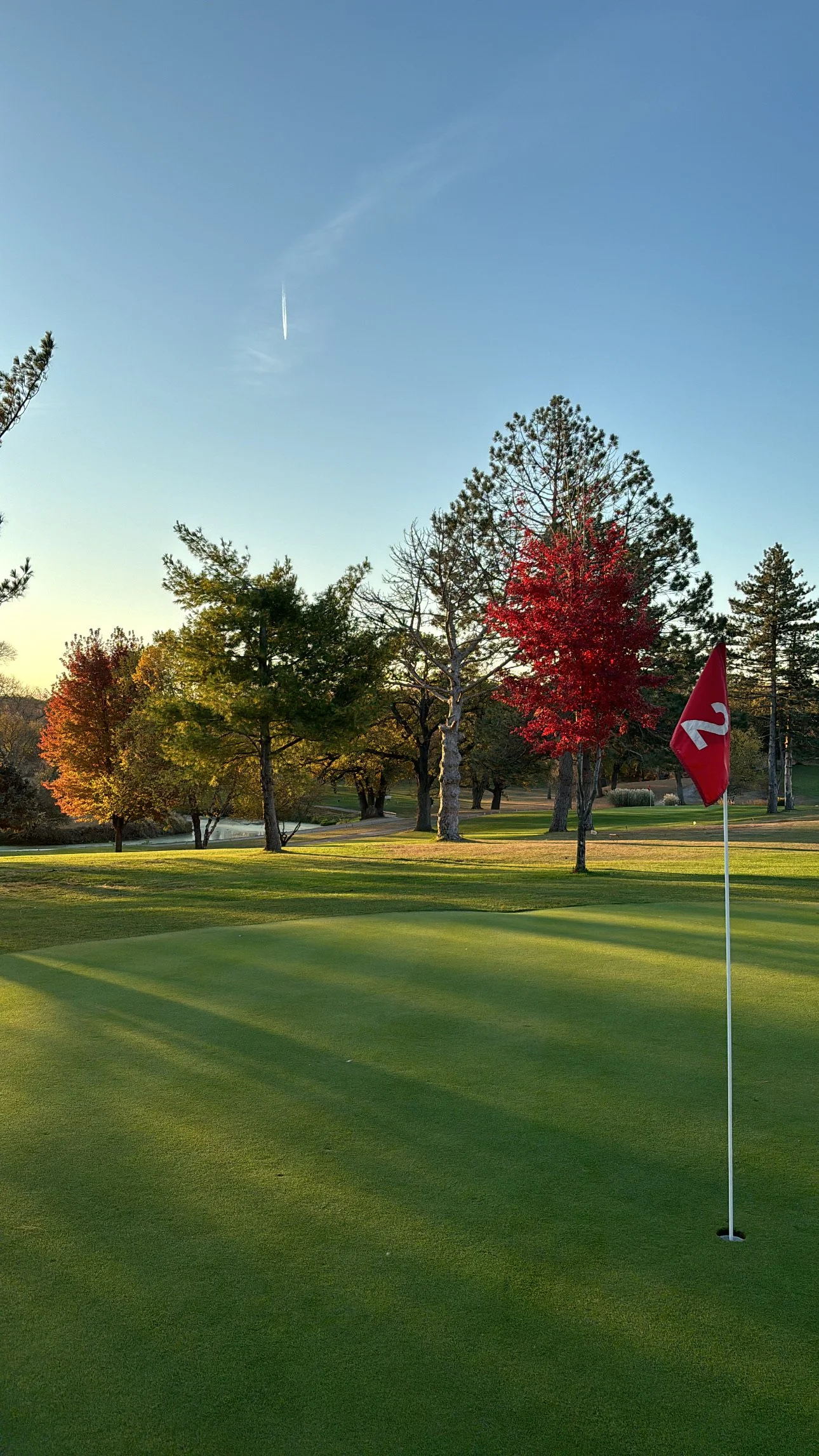 A golf course with green grass and a flagstick marked with the number 2. Trees with autumn leaves are in the background under a blue sky with a jet contrail.