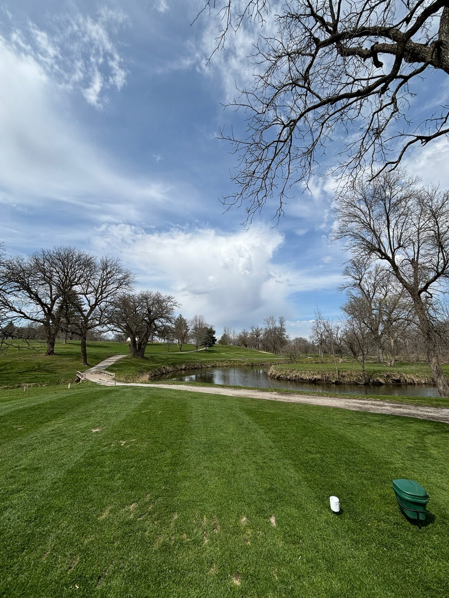 A golf course with a small pond, walking path, and several leafless trees under a partly cloudy sky.