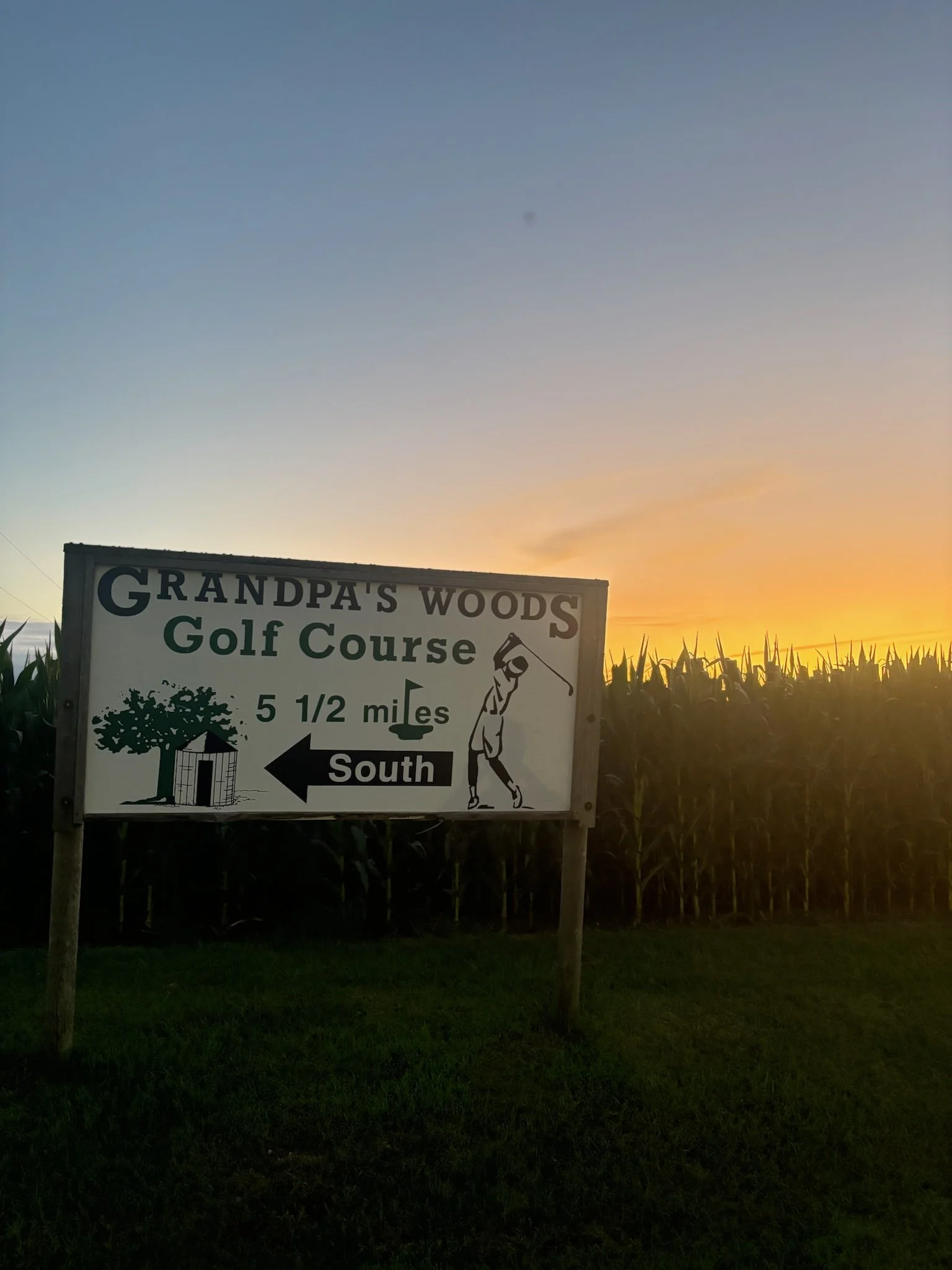 A sign for Grandpa's Woods Golf Course indicating it's 5 and a half miles south, with an illustration of a golfer swinging and a small shed with a tree, set against a sunset sky and a field of crops.