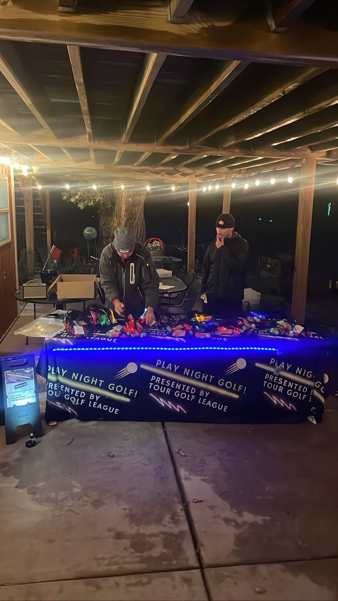 Two men wearing jackets and beanies at a night event under string lights, standing behind a table with a black tablecloth that says "Play Night Golf!" and "Presented by Tour Golf League." one man appears to be managing golf accessories or equipment, while the other man is looking down. There is a large tree in the background and a wooden structure overhead.
