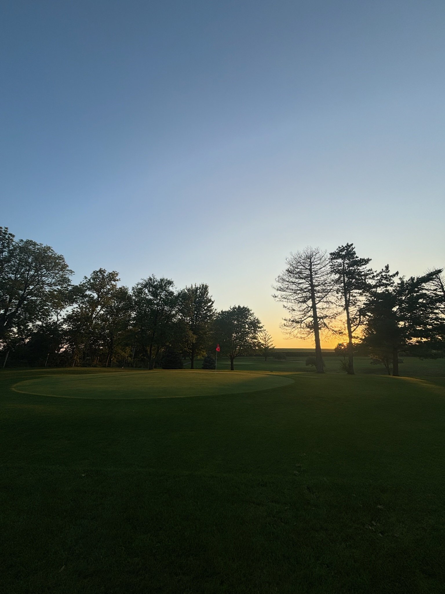 Sunset over a golf course with a flag on the green and trees in the background.