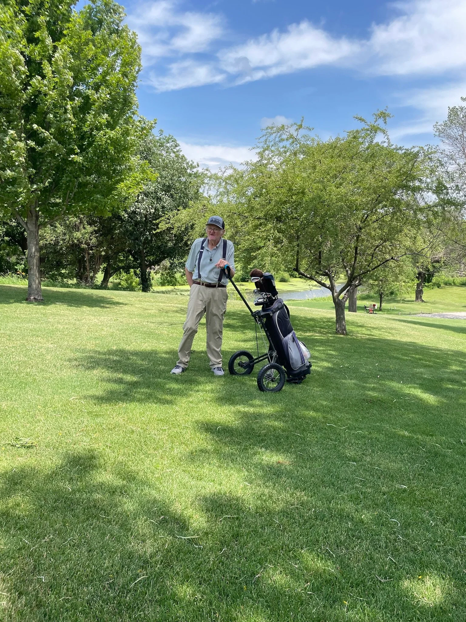 An elderly man stands on a golf course, leaning on a golf club, with a golf bag on a push cart nearby. The background shows green grass, trees, a small pond, and blue sky with some clouds.