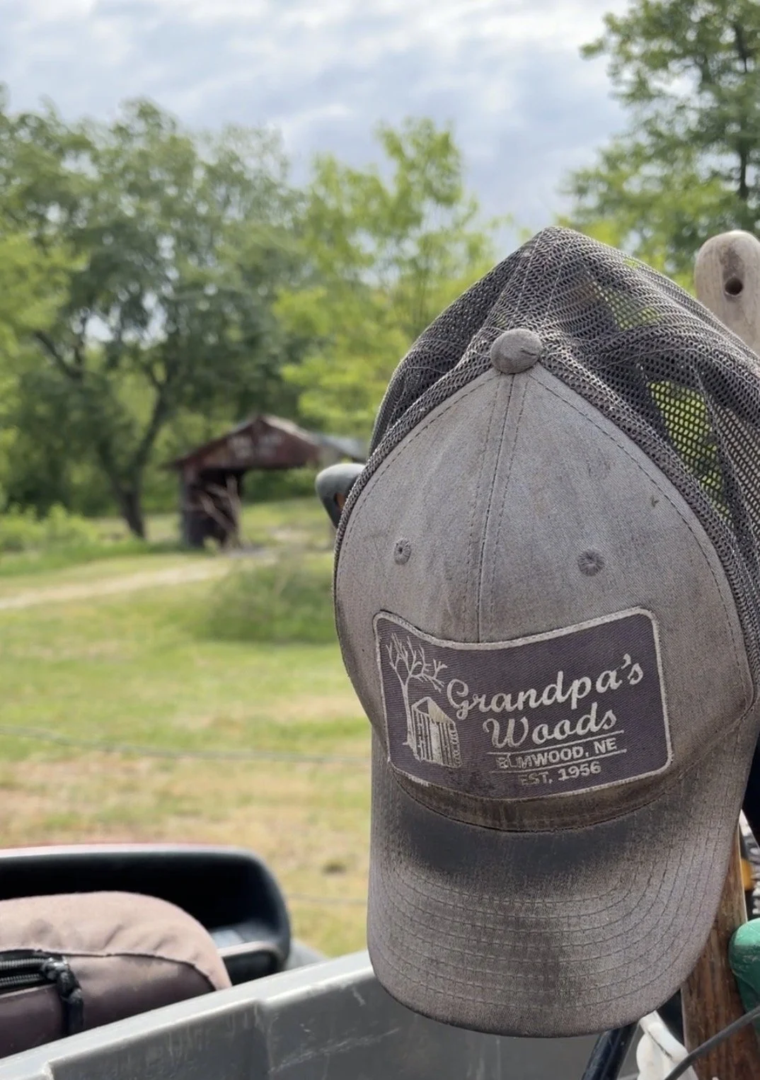 A gray baseball cap with a logo reading 'Grandpa's Woods, Elmwood, NE, Est. 1956' hanging on a wooden fence in a rural outdoor setting with trees and an old shed in the background.
