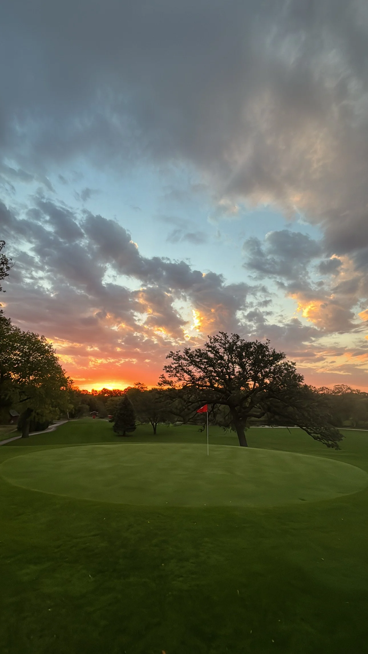 Sunset over a golf course with green grass, trees, and a cloudy sky with hints of orange and yellow near the horizon.