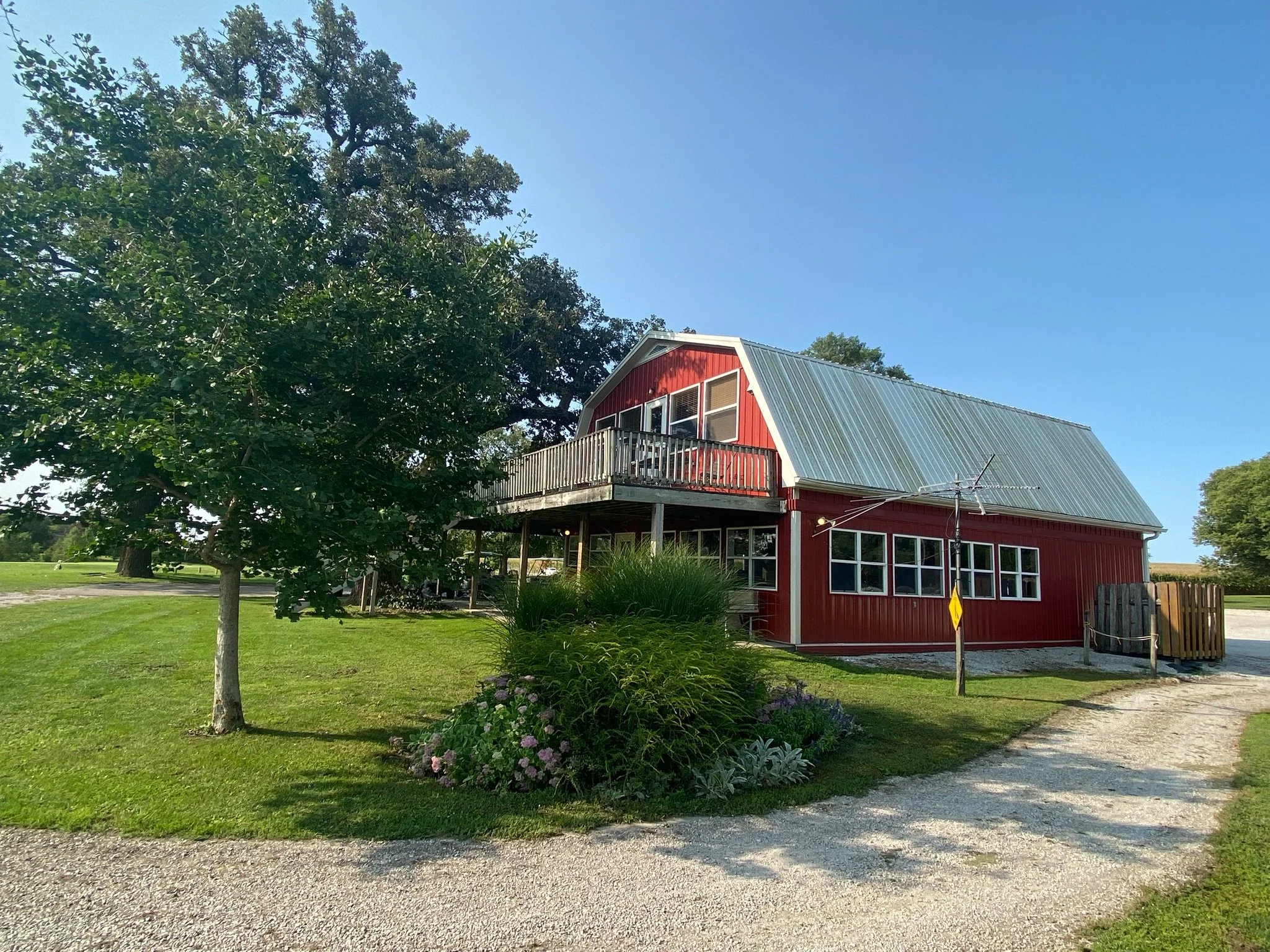 A red barn-style building with a metal roof, windows, and a balcony, situated on a grassy area with trees and a gravel path, under a clear blue sky.