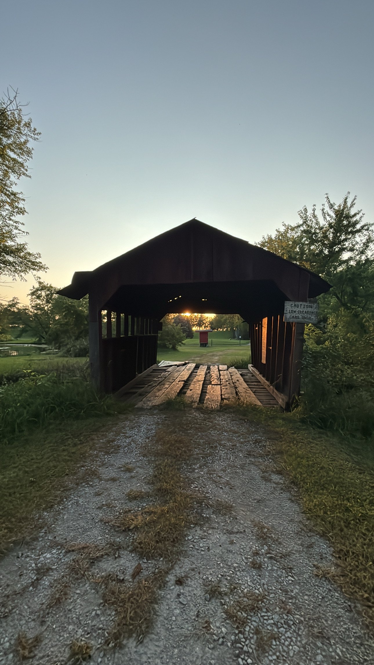 Sunset behind a small covered wooden bridge over a gravel path in a park, surrounded by trees and grass.