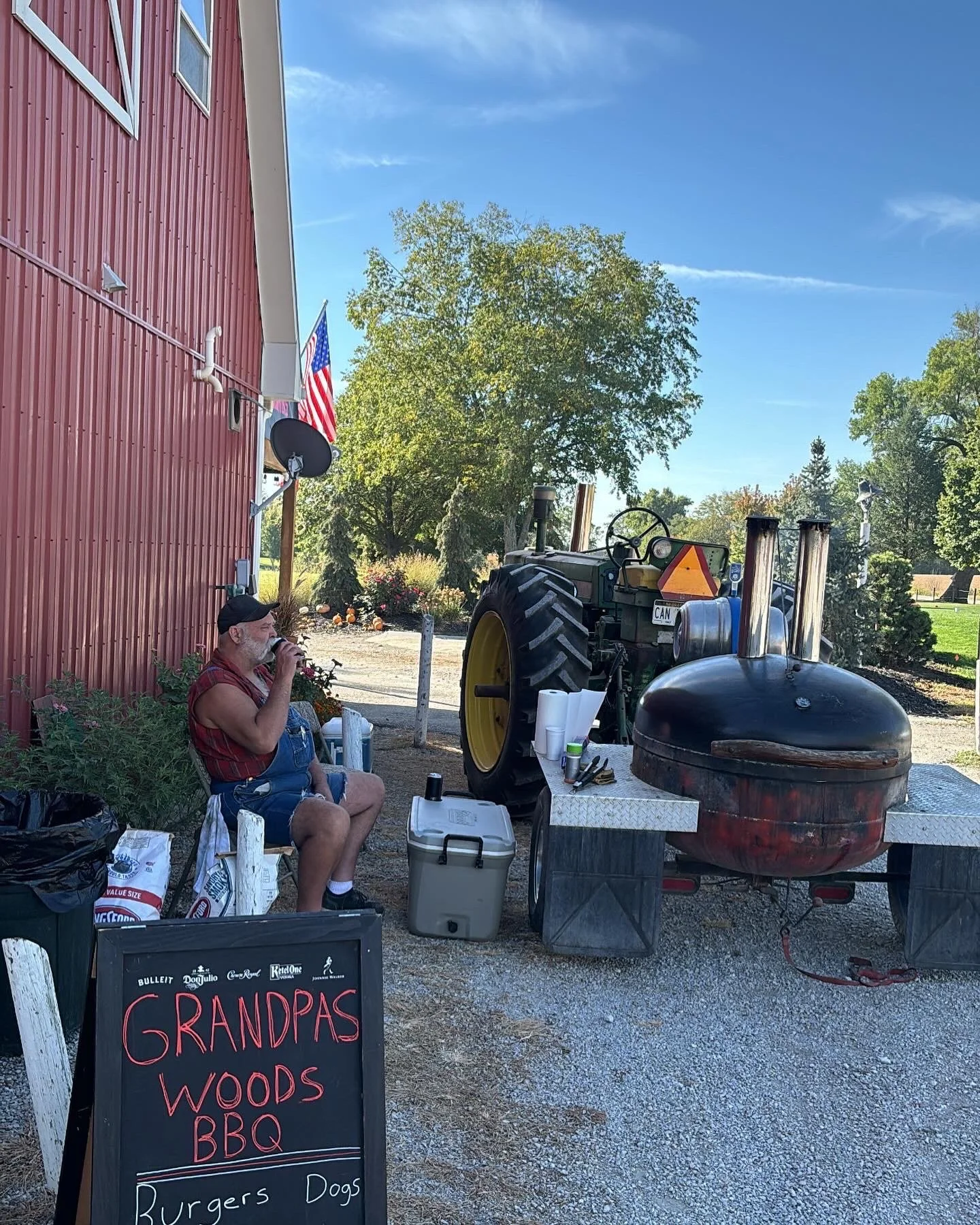 A man with a beard and glasses sitting outside, smoking a pipe, next to a red barn with American flags, a tractor, and a barbecue grill, with a chalkboard sign advertising food.