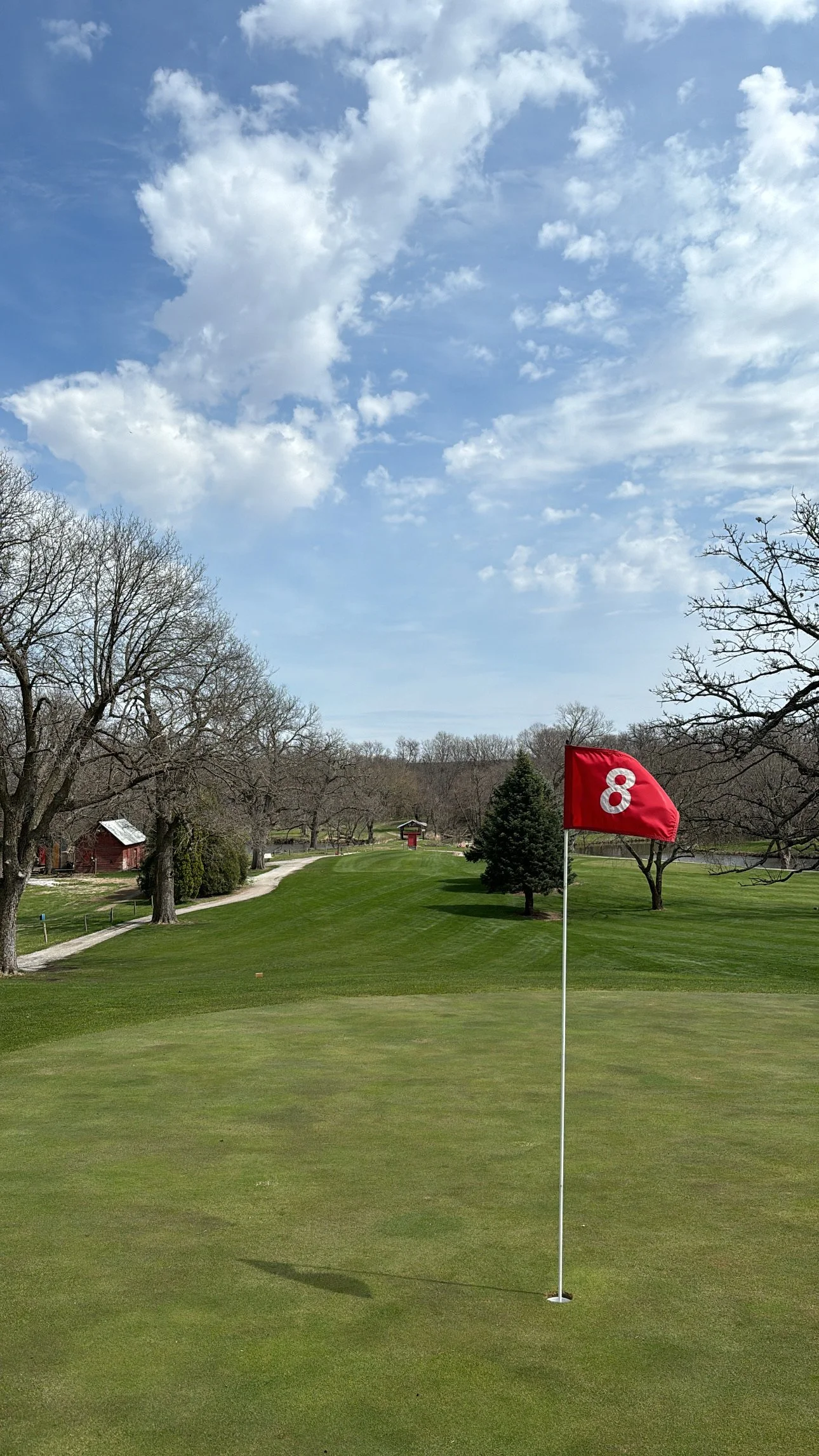 A golf course, putting green, red flag with the number 8, trees, pathway, and a small building under a partly cloudy sky.