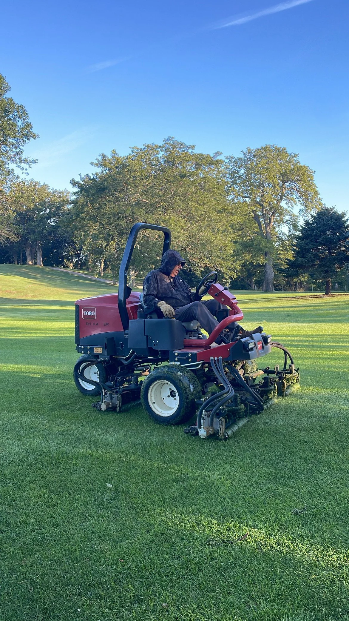 Person wearing a black hooded jacket and gloves operating a red riding lawn mower on a grassy area. There are trees and a clear blue sky in the background.