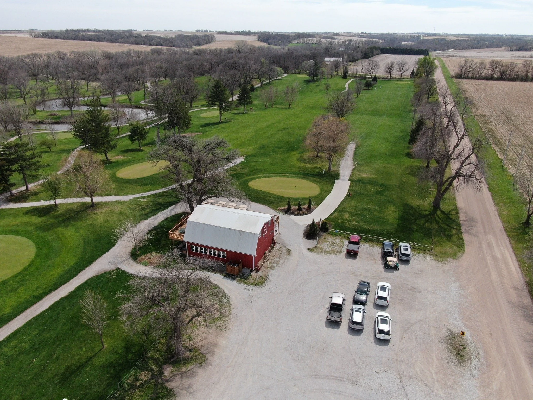Aerial view of a golf course with green grass, trees, a small pond, and surrounding farmland. There is a red building with a white roof in the foreground and parked cars nearby.